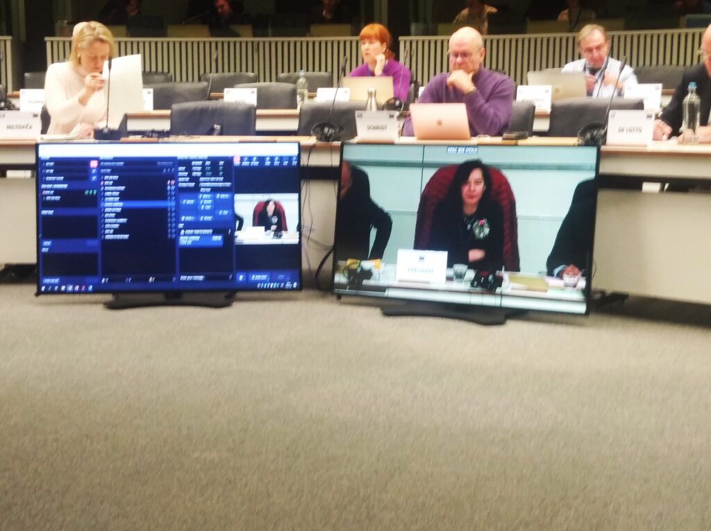 A photo of a group of people at a hearing listening and sitting behind TV monitors